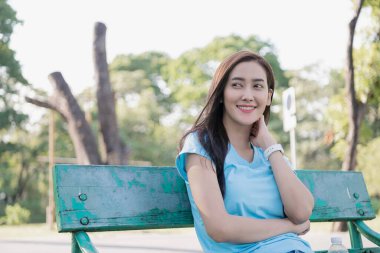 Parktaki bankta oturan bir kadın, güneşli bir günde gülümseyen ve huzurlu ortamın tadını çıkaran bir kadın..