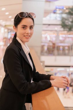 Güneş gözlüklü genç bir iş kadını modern bir alışveriş merkezi ortamında kendinden emin bir şekilde gülümsüyor..