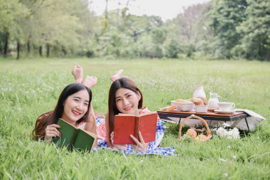 Parkta güneşli bir günün tadını çıkaran iki arkadaş kitap okuyor ve doğanın güzellikleriyle çevrili kahkahalarını paylaşıyorlar..