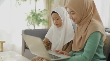 İslami öğrenme konsepti 4k çözünürlük. Asyalı Müslüman anne kızıyla aynı odada oturuyor ve Kuran okumak için dizüstü bilgisayar kullanıyor..