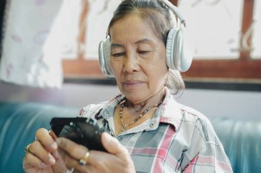 Yaşlı bir kadın akıllı telefonuyla müzik dinlerken kulaklık takıyor ve teknolojinin günlük yaşamdaki etkisini vurguluyor..