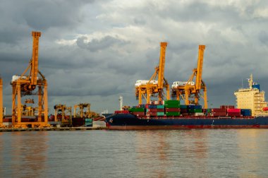 Limana yanaşmış bir kargo gemisi bulutlu bir gökyüzüne doğru yükselen vinçlerle çevrili, endüstriyel taşımacılık sergiliyor..