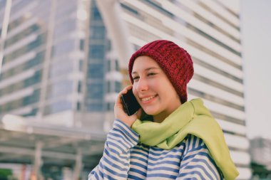 Genç bir kadın, şehir ortamında telefonuyla konuşurken, rahat bir şapka ve eşarp takarak gülümsüyor..