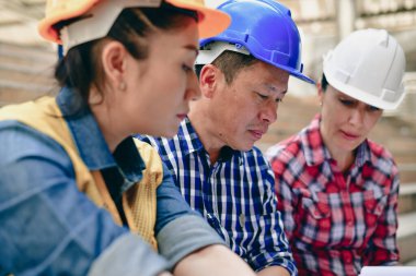 Üç inşaat işçisi bir inşaat sahasında proje planları üzerinde çalışıyor. Başlıkları ve odaklanmış ifadeleri var..