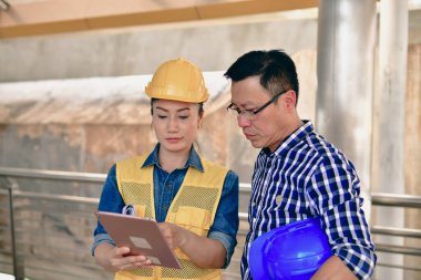İki inşaat işçisi proje detaylarını tartışırken inşaat alanındaki bir tabletin planlarını gözden geçiriyorlar..