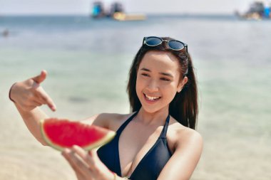 Neşeli bir kadın kumsalda bir dilim karpuzun tadını çıkarır, güneşin altında mutluluk saçar..