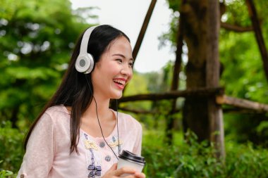 Genç bir kadın, dışarıda kulaklık takıp, yeşil bir ortamda neşe saçarak kahvenin tadını çıkarıyor..