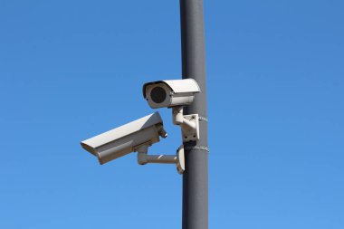 İki küçük sahte modern beyaz devre kamera CCTV güvenlik kameraları zıt yönlere bakıyor koyu gri metal direğe monte edilmiş açık mavi gökyüzü arka planında yerel inşaat alanında