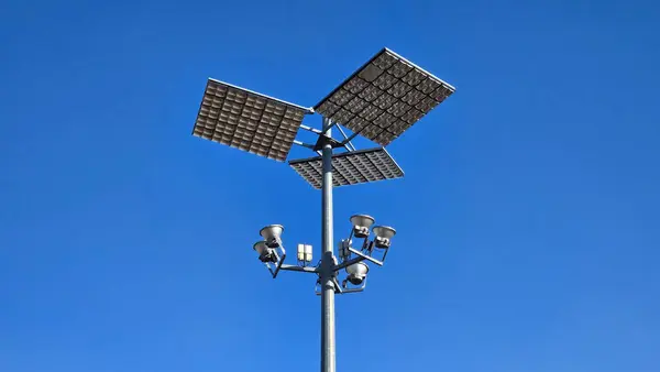 Massive strong light grey metal pole holding modern LED street light reflectors with sensors pointed towards three large reflective panels on clear blue sky background
