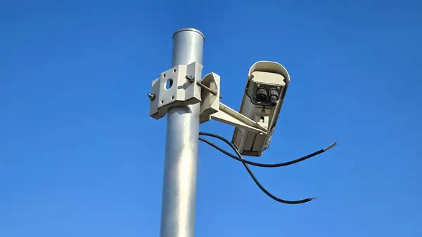Newly installed large white old style closed circuit TV CCTV surveillance camera mounted on strong grey metal pole with large metal mount waiting to be connected with two loose black electrical wires on clear blue sky background