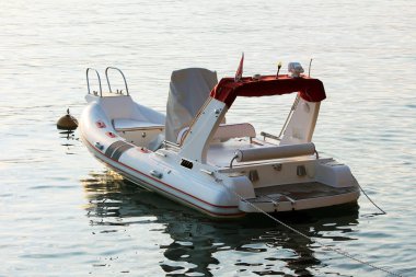 Modern white inflatable flatbeat plastic PVC boat with leather seats and large central steering column anchored in local harbour surrounded with calm blue sea on warm sunny summer day at sunset