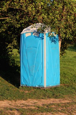 Portatif uniseks mavisi, beyaz ekolojik tuvaleti ya da taşınabilir kimyasal tuvaleti yerel inşaat sahasının kenarına yerleştirilmiş yoğun ağaçların altında sıcak yaz gününde kesilmiş çimlerle çevrili.