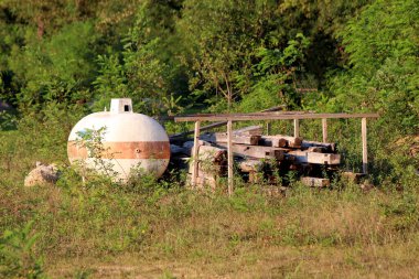 Kahverengi çizgili yıpranmış küresel beyaz tank istiflenmiş ahşap kirişler ve uzun çimenler ve güneşli, aşırı gelişmiş bir tarlada yabani çalılarla çevrili basit bir ahşap çerçeve yanında duruyor..