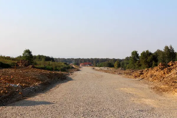 İnşaat halindeki geniş çakıl yolu uzak bir eve doğru uzanıyor, toprak yığınları ve yeşil ağaçlarla çevrili, açık güneşli bir günde geleceğin yolunu gösteren ahşap kazıklar var..