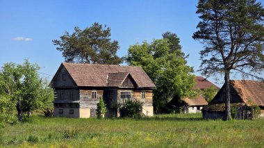 Eski ek binalar ve kır çiçekleri ile çevrili, kiremitli çatılı ahşap çiftlik evi, sakin bir kırsal kesimde berrak mavi bir gökyüzünün altında, ağaçlar ve çimenlerin arasında duruyor..