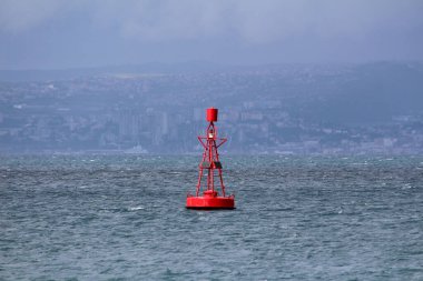Güneş panelleri ve kafes kapaklı parlak kırmızı navigasyon şamandırası dalgalı bir denizde yüzer, uzak şehir silueti sisli ve alçak bulutların arasından görünür..