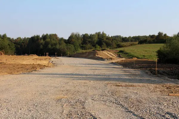 İnşaat halindeki geniş çakıllı yol, toprak yığınları, çim tarlaları ve uzak ormanlarla çevrili kırsal alanlara uzanır. Geleceğe giden yolu gösteren ahşap kazıklarla çevrili..