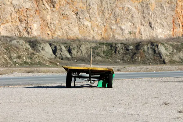 Yıpranmış sarı düz karavanlı, iki tekerleği ve açık çerçeveli, kaldırımlı bir yolun yakınındaki çakılların üzerinde, parlak güneş ışığı altında, dik bir kayalık yamaçla desteklenmiş..