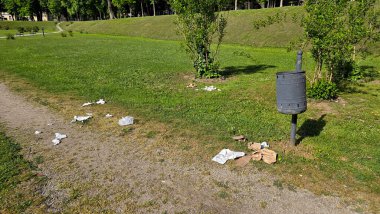 Çevresi ağaçlar ve çalılarla çevrili yeşil çimenlerin üzerinde duran dağınık çöp tenekeleri ile park yolu, halka açık yeşil bir alanda çevresel ihmalkarlığı vurguluyor.