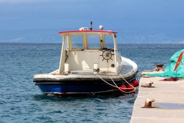 Kırmızı bukleli sıkıştırılmış beyaz ve mavi römorkör, bulutlu bir gökyüzünün altında kırmızı şamandıralı sakin deniz suyunda yüzen beton bir rıhtıma bağlıdır..