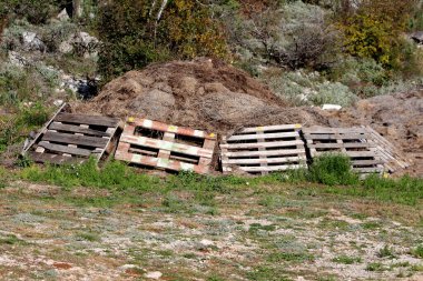 Dört eski ahşap palet büyük bir gübre yığınına yaslanır saman ve organik malzeme, kayalık, aşırı gelişmiş kırsal arazi vahşi bitkiler ve çalılar ile.