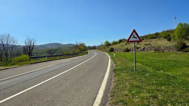 Çimenli yamaçlar ve ormanla çevrili eğimli asfalt yol, bir üçgen geyik geçiş işareti ile işaretlenmiş; rüzgar türbinleri ve açık mavi bir gökyüzünün altındaki yeşil tepelerde görünen bir baz istasyonu