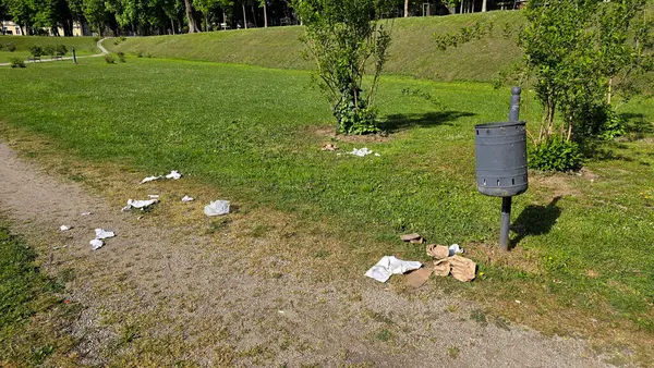 Çevresi ağaçlar ve çalılarla çevrili yeşil çimenlerin üzerinde duran dağınık çöp tenekeleri ile park yolu, halka açık yeşil bir alanda çevresel ihmalkarlığı vurguluyor.