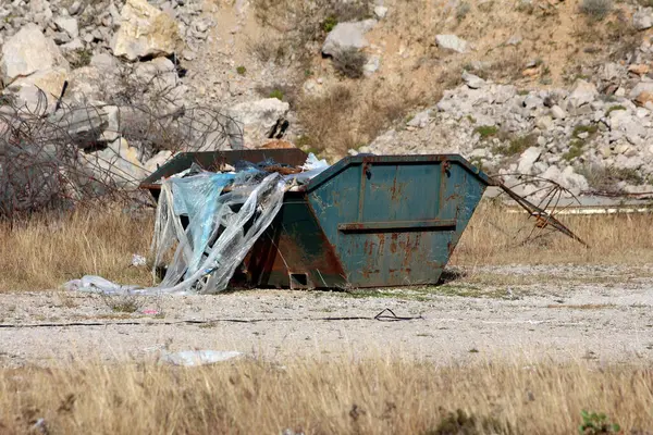 Plastik çarşaf ve molozlarla dolu büyük paslı yeşil çöp konteynırları kuru, çimenli bir alanda duruyor. Dağılmış kayalar ve bükülmüş metallerle endüstriyel bir alanda atık yönetimini vurguluyor..