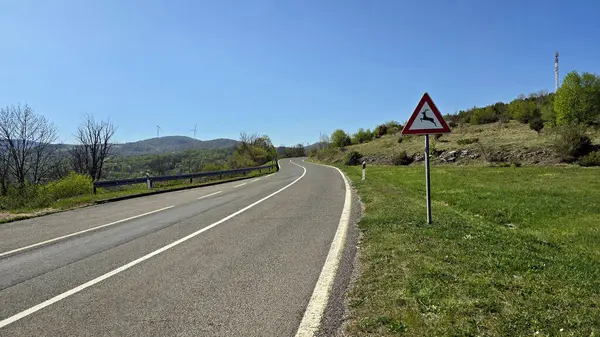 Çimenli yamaçlar ve ormanla çevrili eğimli asfalt yol, bir üçgen geyik geçiş işareti ile işaretlenmiş; rüzgar türbinleri ve açık mavi bir gökyüzünün altındaki yeşil tepelerde görünen bir baz istasyonu