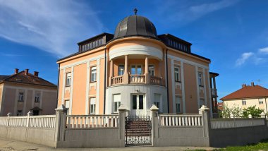 Geniş merkezi kubbeli zarif tarihi bir villa sütunlarla desteklenen yuvarlak bir balkon ve şeftali rengi cephe beyaz kapılı bir çitin arkasında duruyor, sakin bir yerleşim bölgesinde açık mavi bir gökyüzüne karşı kurulmuş.