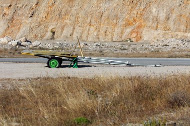 Yeşil tekerlek ve metal çerçeveli eskimiş karavan kaldırımlı bir yolda terk edilmiş, dik, kayalık bir taş ocağı duvarı tarafından desteklenmiş, parlak güneş ışığı altında, kırsal sanayi ve çürümeyi çağrıştırıyor.