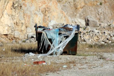 Plastik çarşaf ve molozlarla dolu büyük paslı yeşil çöp konteynırları kuru, çimenli bir alanda duruyor. Dağılmış kayalar ve bükülmüş metallerle endüstriyel bir alanda atık yönetimini vurguluyor.