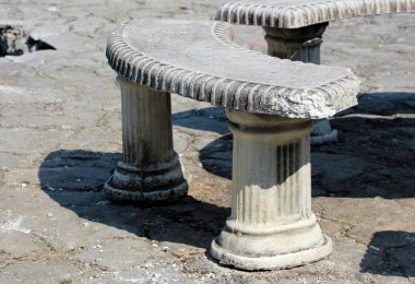 Süslü flüt sütunlu yıpranmış bir taş bank asfalt bir yüzey üzerinde duruyor, yaş ve yontma belirtileri gösteriyor, klasik bahçe tasarımı ve açık hava rahatlama çağrıştırıyor