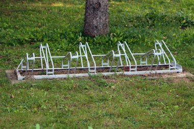 Birçok yuvası olan beyaz metal bir bisiklet parktaki çimenli bir çimenlikte duruyor, yeşil yapraklarla çevrili ve bir ağaç tarafından gölgelenmiş, açık havada bisikletçiler tarafından kullanılmaya hazır.