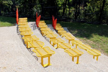 Yuvarlak sırtlı, parlak sarı metal koltuklar ve ağ yüzeyleri, güneş ışığı altında çimlerin ve ağaçların bulunduğu çakıl yüzeyinde, kırmızı korkuluklarla ayrılmış eğimli bir açık hava amfi tiyatrosunda düzenlenir.