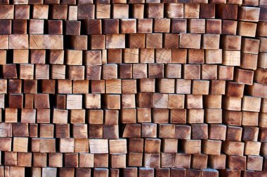 Kare şeklinde büyük bir ahşap kiriş yığını, tahılın ucu dışa dönük, doğal ahşap desenler, çatlaklar ve parlak ışık altında tekrarlayan geometrik desen içinde sıcak kahverengi tonlar sergiliyor.