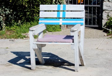 Mavi çizgili ve lavanta koltuklu sağlam beyaz bir tahta koltuk güneşli beton bir verandada oturur, koyu bir gölge bırakır, arka planda yeşil yapraklar ve siyah metal bir kapı vardır.