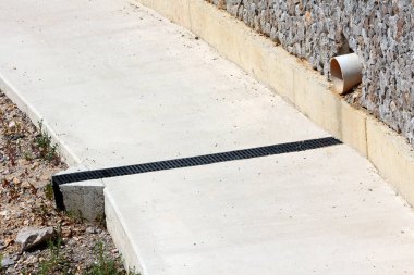Hafif beton drenaj kanalı, beyaz bir akış borusu ve siyah plastik ızgara, eğimli bir sitedeki fırtına suyunu yönetmek üzere tasarlanmış taş bir istinat duvarının yanında çalışır.