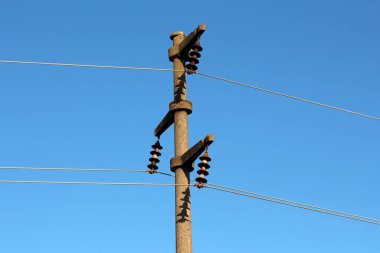 Seramik disk izolatörleri ve elektrik hatları olan uzun beton elektrik direği açık mavi gökyüzüne uzanır ve parlak güneş ışığında keskin gölgeler bırakır.