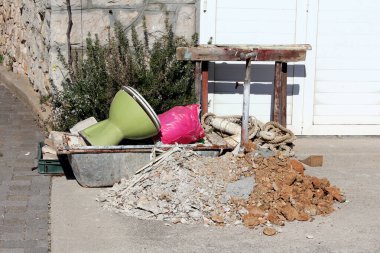 Yeşil seramik tuvaleti, pembe çantası, ipleri, ahşabı ve kırık betonu olan inşaat atığı, taş duvarın ve otlar ve güneş ışığıyla çevrili beyaz kapıların yanındaki kaldırımda duruyor.
