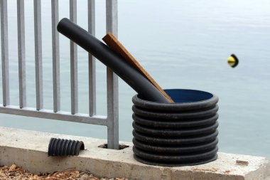 Siyah eğimli boru ve ahşap tahta büyük plastik bir halka içinde metal bir çitin yanında beton bir kenarda, arka planda durgun suda yüzen bir şamandıra ile dinleniyor.