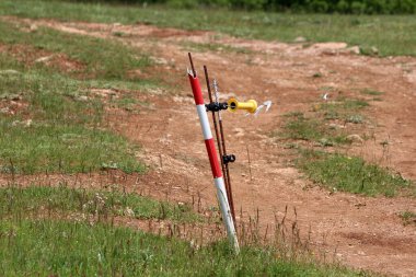 Sarı ve siyah yalıtkan ve metal çubuklu kırmızı ve beyaz çizgili elektrikli çit direği yeşil çimenler ve kır çiçekleriyle çevrili toprak çiftlik yolu boyunca bir kabloyu destekliyor.