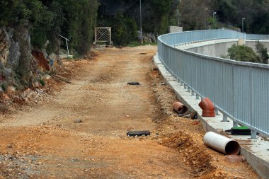 Bitmemiş çakıl yolu uzun metal bir korkuluk boyunca kıvrımlar, açıkta kalan drenaj boruları, ızgaralar ve kayalık kesim yamaç, ağaçlar ve çalılarla çevrili küçük bir kapıda sona erer