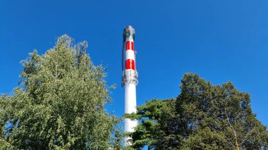 Kırmızı ve beyaz çizgilerle boyanmış uzun sanayi bacası yapraklı ağaçların üzerinde yükseliyor, tepesine yakın iskele ve ağlarla aydınlık, güneşli bir günde mavi gökyüzüne karşı kuruluyor..