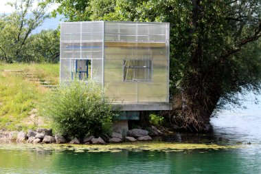 Yarı saydam polikarbonat duvarları ve açık pencereleri olan küçük modern sığınak güneşli bir günde sakin göle yansıyan yemyeşil ve kayalarla çevrili suyun üzerinde duruyor.