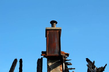 Kömürleşmiş ahşap kirişler ve tuğlalar üzerinde yükselen paslı metal baca, parlak mavi gökyüzüne karşı dizilmiş, ateş tarafından tahrip edilmiş bir binanın kalıntılarını işaret ediyor.