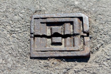 Katmanlı geometrik tasarımı olan eski paslı kare rögar kapağı sert asfalta yerleştirilmiştir. Güçlü güneş ışığıyla yıpranma, aşınma ve kentsel dokuyu gösterir.