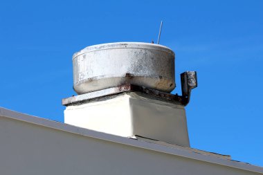 Pas ve lekeli büyük yuvarlak metal baca kapağı düz beyaz bir çatının tepesinde, kare bir zemine monte edilmiş, güçlü mavi bir gökyüzüne karşı keskin bir şekilde duruyor.