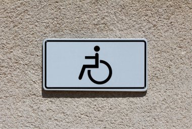 Beyaz arka planda siyah sembollü basit tekerlekli sandalye erişim tabelası kaba bej bir duvara monte edilmiştir. Bu da engelliler için engelsiz erişimi gösterir.
