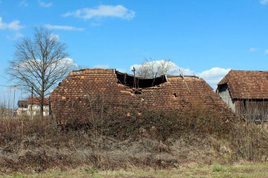 Çökmüş kırmızı kiremit çatılı eski bir kırsal bina ve aşırı gelişmiş bitki örtüsü, kuru çimenler ve yapraksız çalılarla çevrili, dağınık bulutlu mavi bir gökyüzünün altında